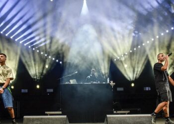 Two performers with microphones stand on stage at Fiesta Voio 2026, illuminated by bright white spotlights while a DJ is visible behind them.