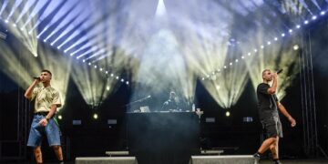 Two performers with microphones stand on stage at Fiesta Voio 2026, illuminated by bright white spotlights while a DJ is visible behind them.