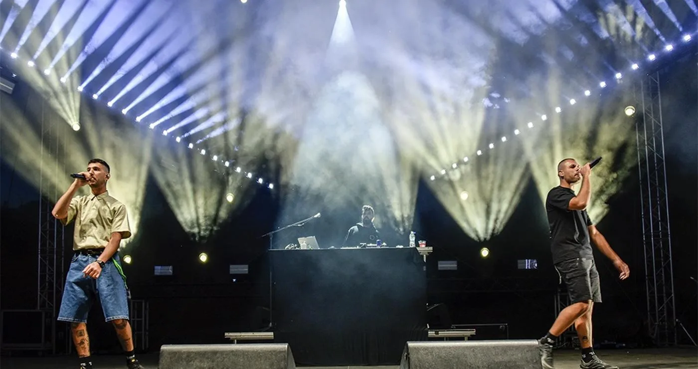 Two performers with microphones stand on stage at Fiesta Voio 2026, illuminated by bright white spotlights while a DJ is visible behind them.