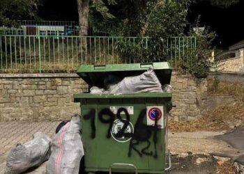 Ένας πράσινος μεταλλικός κάδος για οικοδομικά απόβλητα, γεμάτος με μπάζα και τσουβάλια, βρίσκεται σε έναν δρόμο με λιθόστρωτο στην Φλώρινα.