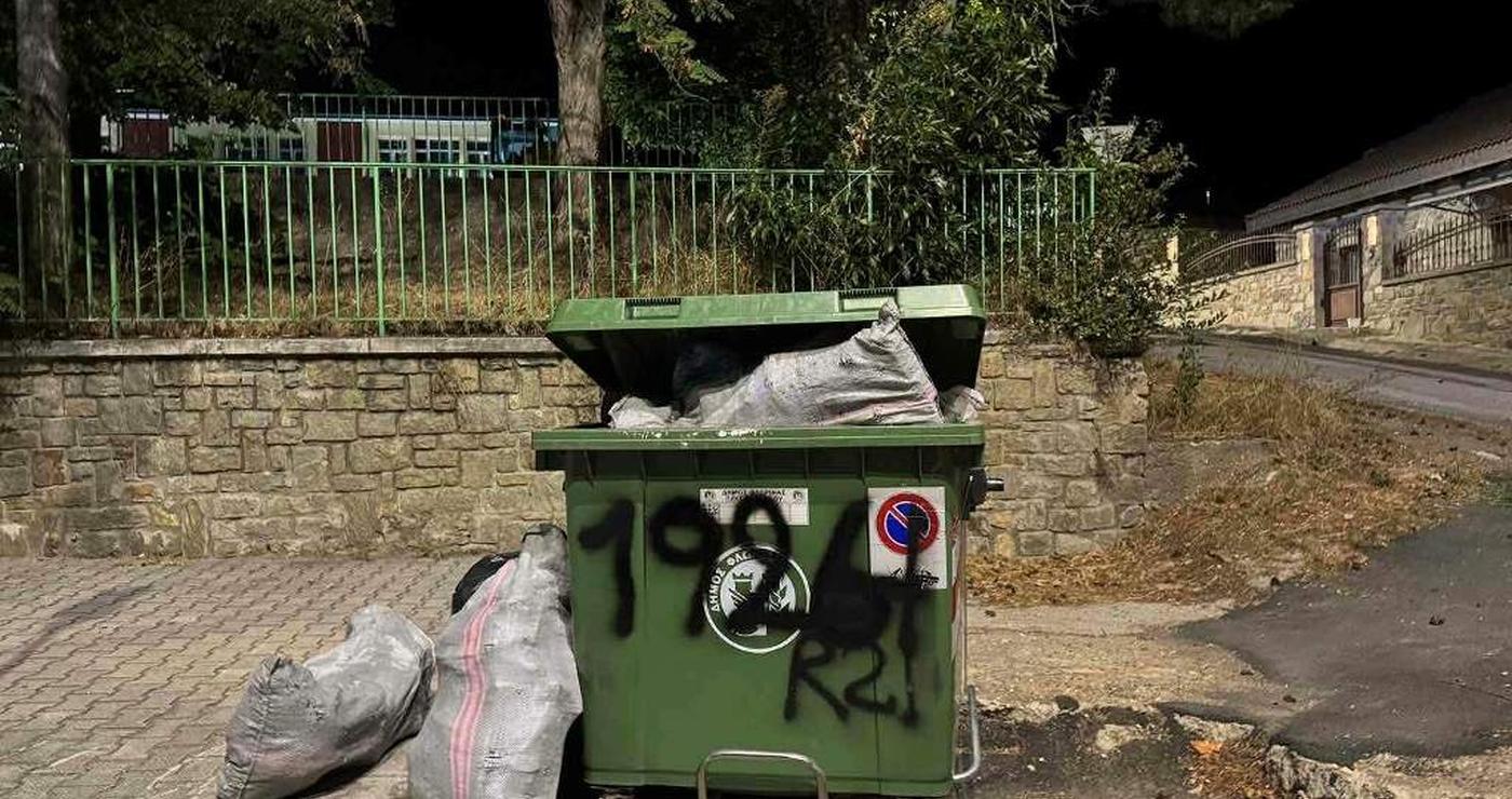 Ένας πράσινος μεταλλικός κάδος για οικοδομικά απόβλητα, γεμάτος με μπάζα και τσουβάλια, βρίσκεται σε έναν δρόμο με λιθόστρωτο στην Φλώρινα.