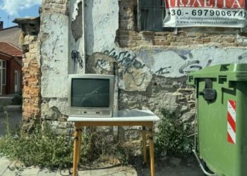 Ένα παλιό τηλεοπτικό σετ στέκεται πάνω σε ένα ξύλινο τραπέζι δίπλα σε έναν πράσινο κάδο ογκωδών απορριμμάτων σε έναν δρόμο της Φλώρινας, με ένα παλιό κτίριο και μια κόκκινη διαφήμιση στο φόντο.
