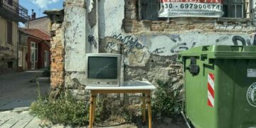 Ένα παλιό τηλεοπτικό σετ στέκεται πάνω σε ένα ξύλινο τραπέζι δίπλα σε έναν πράσινο κάδο ογκωδών απορριμμάτων σε έναν δρόμο της Φλώρινας, με ένα παλιό κτίριο και μια κόκκινη διαφήμιση στο φόντο.