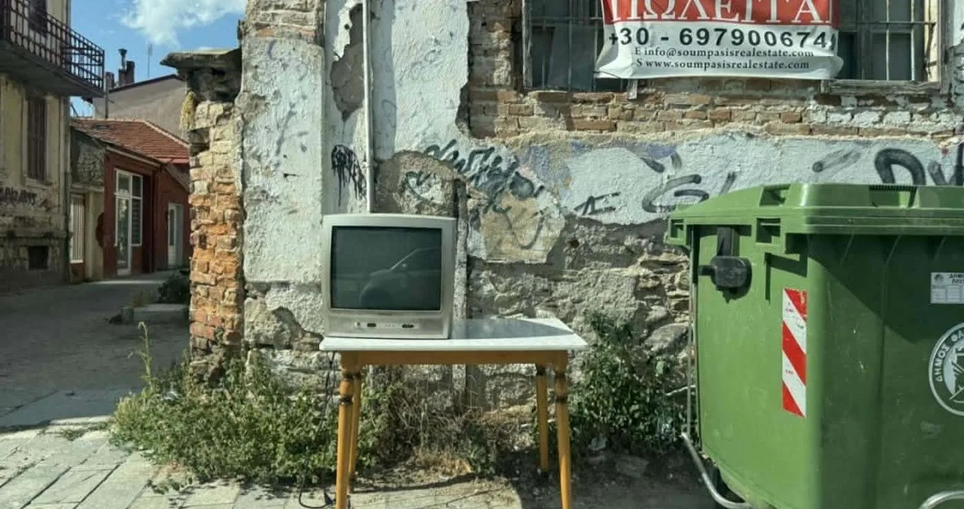 Ένα παλιό τηλεοπτικό σετ στέκεται πάνω σε ένα ξύλινο τραπέζι δίπλα σε έναν πράσινο κάδο ογκωδών απορριμμάτων σε έναν δρόμο της Φλώρινας, με ένα παλιό κτίριο και μια κόκκινη διαφήμιση στο φόντο.