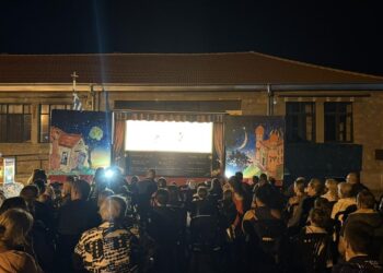 Ένα κοινό παρακολουθεί με ενθουσιασμό μια παράσταση Θεάτρου Σκιών Σκλήθρο σε έναν φωτισμένο προαύλιο χώρο σχολείου το βράδυ.