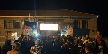 Ένα κοινό παρακολουθεί με ενθουσιασμό μια παράσταση Θεάτρου Σκιών Σκλήθρο σε έναν φωτισμένο προαύλιο χώρο σχολείου το βράδυ.