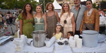 Μια ομάδα ανθρώπων στέκεται πίσω από έναν πάγκο με ποτήρια και κατσαρόλες, προσφέροντας Πατρίδων Γεύσεις σε μια εκδήλωση στο Νέο Πάρκο της Φλώρινας.