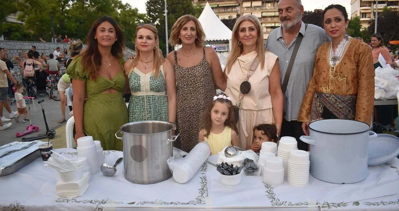 Μια ομάδα ανθρώπων στέκεται πίσω από έναν πάγκο με ποτήρια και κατσαρόλες, προσφέροντας Πατρίδων Γεύσεις σε μια εκδήλωση στο Νέο Πάρκο της Φλώρινας.