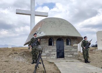 Δύο Έλληνες στρατιώτες στέκονται σε προσοχή μπροστά από το μνημείο στην Αννίτσα των Γρεβενών, ένα πέτρινο εκκλησάκι με έναν μεγάλο σταυρό στο ύψος, σε μια ηλιόλουστη μέρα.