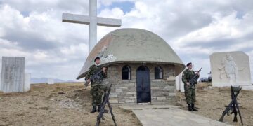 Δύο Έλληνες στρατιώτες στέκονται σε προσοχή μπροστά από το μνημείο στην Αννίτσα των Γρεβενών, ένα πέτρινο εκκλησάκι με έναν μεγάλο σταυρό στο ύψος, σε μια ηλιόλουστη μέρα.