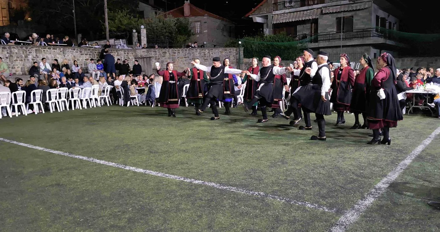Μια ομάδα χορευτών από τους Γρεβενιώτες Κοζάνης χορεύουν σε ένα γήπεδο στην Κλεισούρα, με θεατές να παρακολουθούν το παραδοσιακό γλέντι.