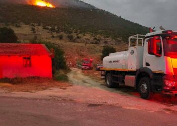A white fire truck stands on a road near a building, with flames and smoke rising from a δασική πυρκαγιά on a hillside in the background.