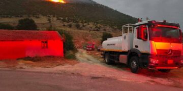 A white fire truck stands on a road near a building, with flames and smoke rising from a δασική πυρκαγιά on a hillside in the background.