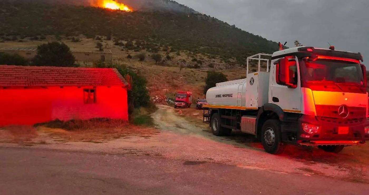 A white fire truck stands on a road near a building, with flames and smoke rising from a δασική πυρκαγιά on a hillside in the background.