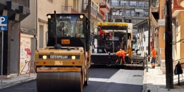 Μια μεγάλη πορτοκαλί κύλινδρος συμπιέζει την καινούργια άσφαλτο σε έναν κεντρικό δρόμο της Φλώρινας, στο πλαίσιο των ασφαλτοστρώσεων.