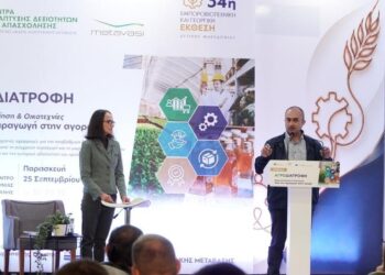 Ένας άνδρας παρουσιαστής μιλάει σε ένα κοινό στην ημερίδα αγροδιατροφής στην Δυτική Μακεδονία, με μια μεγάλη οθόνη πίσω του να δείχνει μια σύνθεση από προϊόντα και γραφήματα.