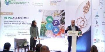 Ένας άνδρας παρουσιαστής μιλάει σε ένα κοινό στην ημερίδα αγροδιατροφής στην Δυτική Μακεδονία, με μια μεγάλη οθόνη πίσω του να δείχνει μια σύνθεση από προϊόντα και γραφήματα.