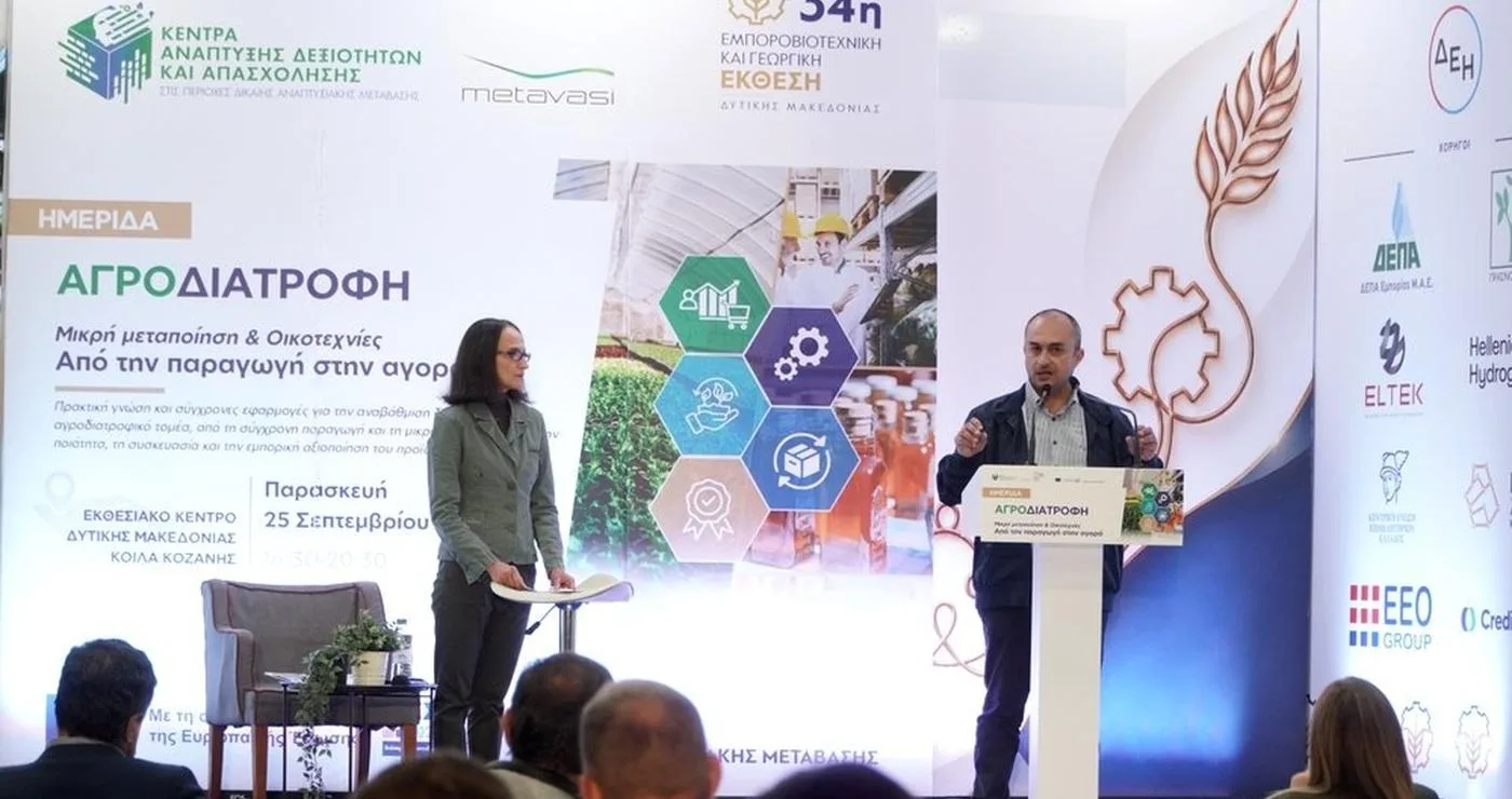 Ένας άνδρας παρουσιαστής μιλάει σε ένα κοινό στην ημερίδα αγροδιατροφής στην Δυτική Μακεδονία, με μια μεγάλη οθόνη πίσω του να δείχνει μια σύνθεση από προϊόντα και γραφήματα.