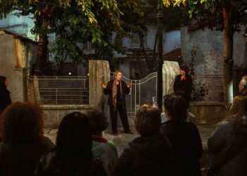 Μια ηθοποιός απευθύνεται σε ένα μικρό κοινό σε έναν εξωτερικό χώρο κατά τη διάρκεια του Κοζάνη περίπατος, με παραδοσιακά σπίτια και πράσινο στο φόντο.